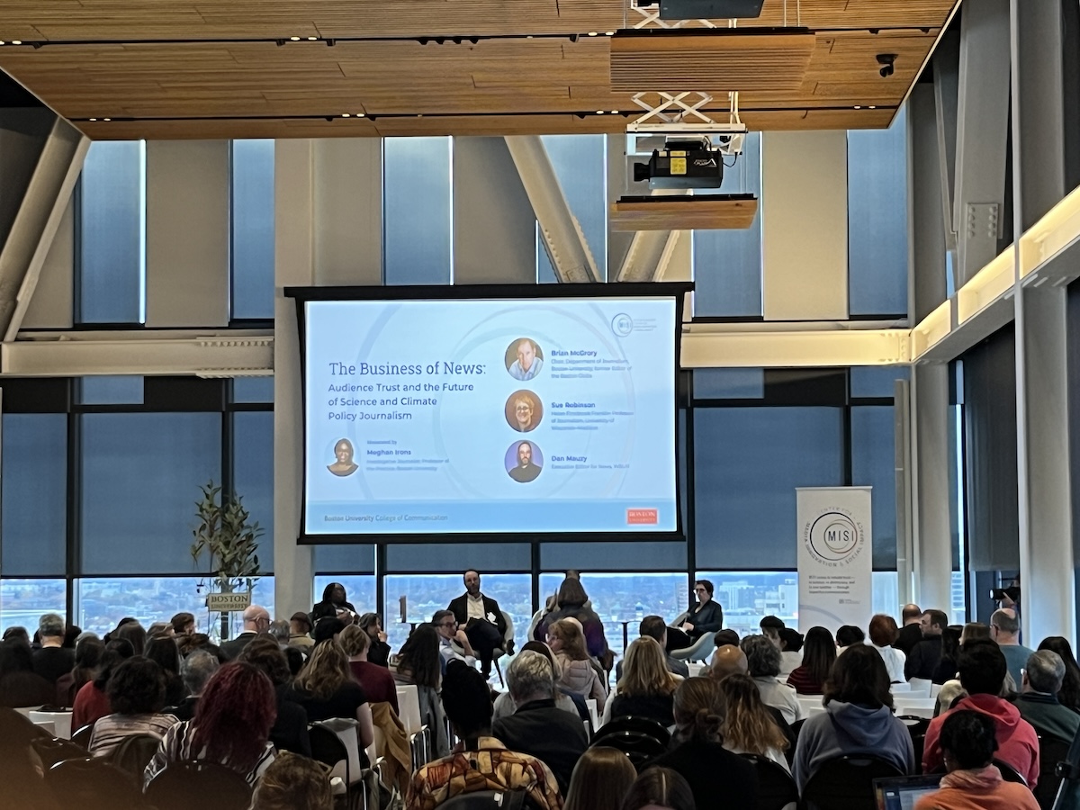photo from behind audience at opening of panel talk, with speakers on stage and slide introducing them on a big projected screen behind