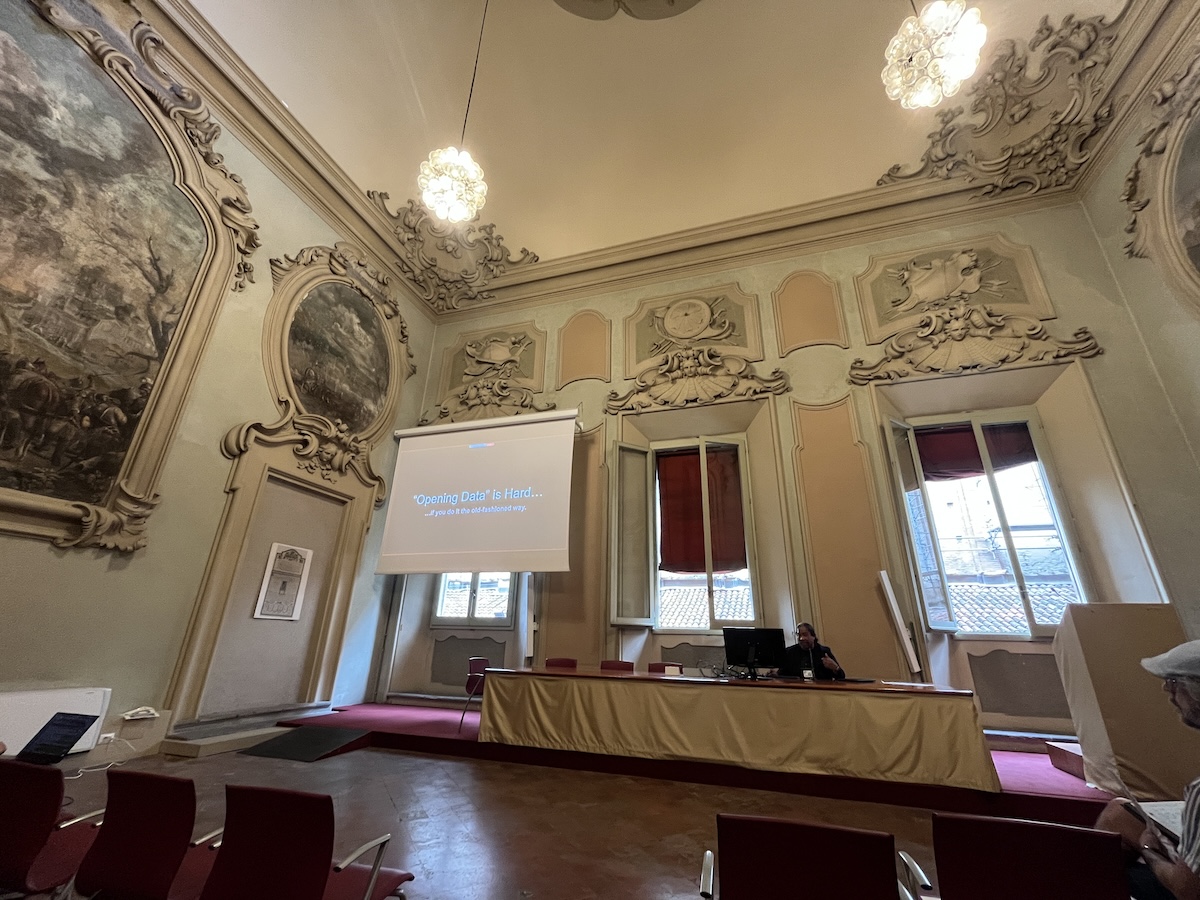 a wide photo of an ornate room with a man sitting up front behind a desk and a projected slide to his right