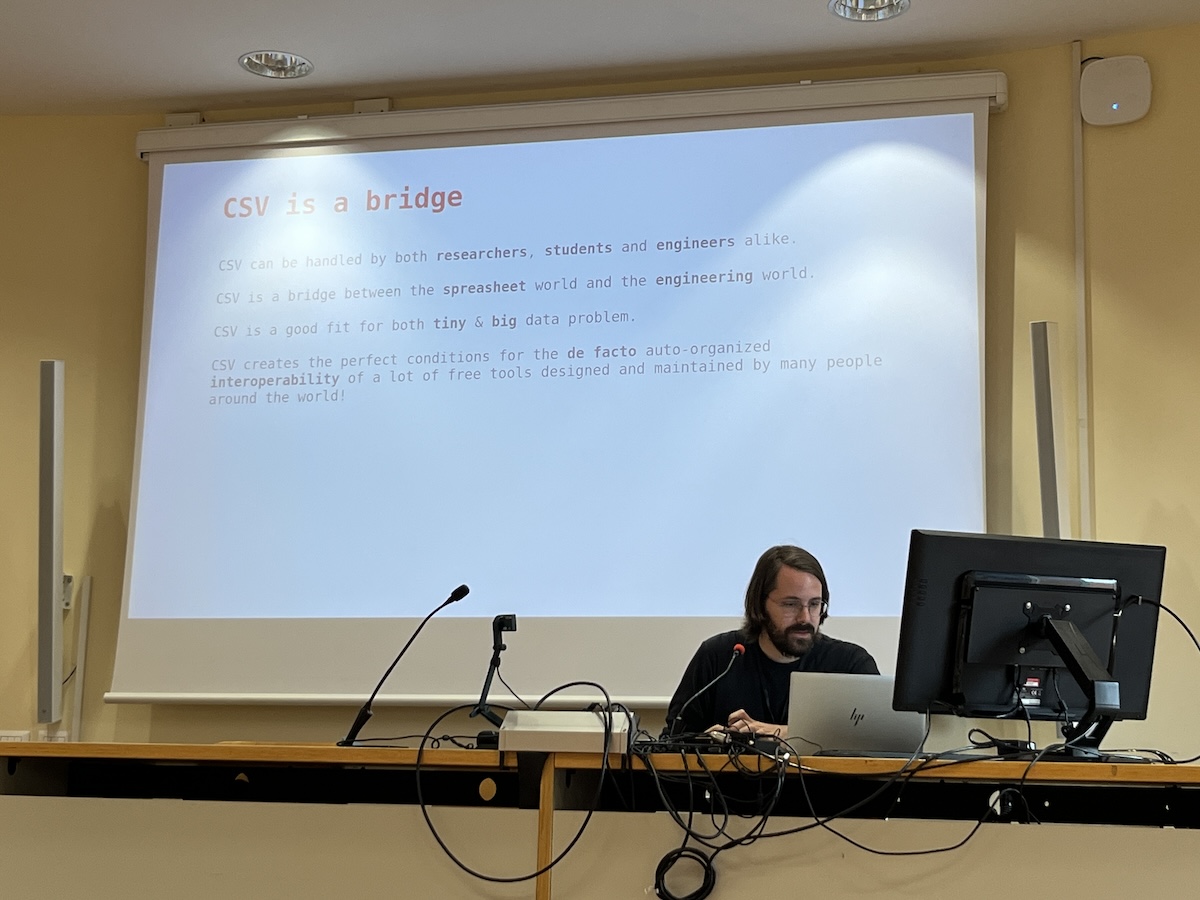 a man with dark hair and a beard sitting at a computer with a large slide about CSV content behind him