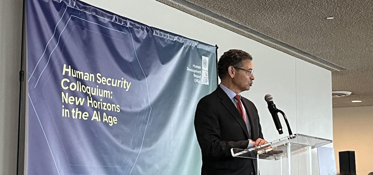 speaking man with short hair standing behind podium in front of banner with event name